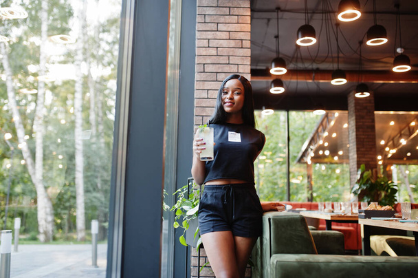 Módní feministka africká Američanka v černém tričku a šortkách, pózuje v restauraci s limonádovým sklem. - Fotografie, Obrázek