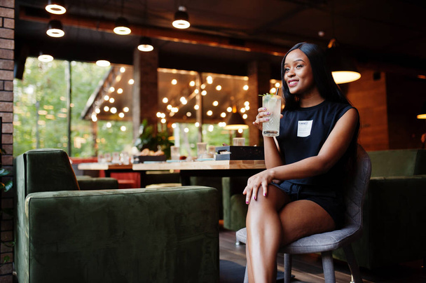 Fashionable feminist african american woman wear in black t-shirt and shorts, posed at restaurant with lemonade glass. - 写真・画像
