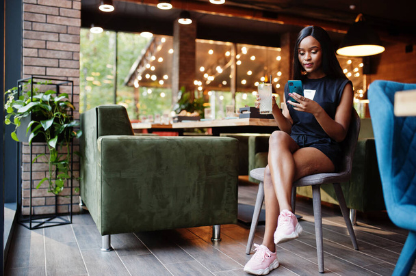 Fashionable feminist african american woman wear in black t-shirt and shorts, posed at restaurant with lemonade glass and mobile phone. - Fotografie, Obrázek