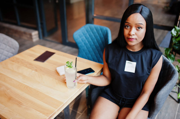 Fashionable feminist african american woman wear in black t-shirt and shorts, posed at restaurant with lemonade glass. - Φωτογραφία, εικόνα