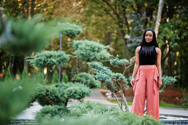 Μοντέρνα αφροαμερικάνικη γυναίκα με ροδακινί παντελόνι και μαύρη μπλούζα ποζάρουν εξωτερική. - Φωτογραφία, εικόνα