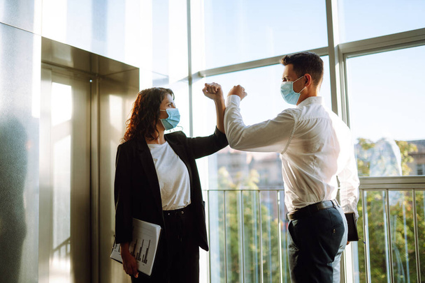 Business colleagues in protective face masks bumping elbows while greeting each other near elevator at modern office. Covid-19. - Foto, immagini