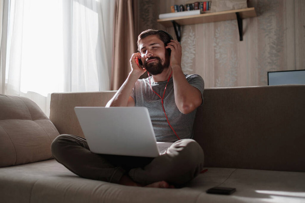 Άνδρας στον καναπέ με ακουστικά και σημειωματάριο στο σπίτι. οι νέοι hipster φορούν ακουστικά σπουδάζουν online παρακολουθώντας webinar podcast σε laptop ακούγοντας μαθήματα εκμάθησης ή μουσικής, μαθαίνοντας έννοια - Φωτογραφία, εικόνα