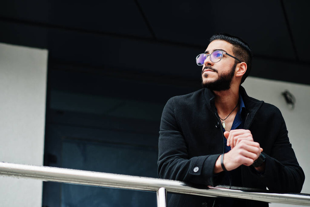 Middle eastern entrepreneur wear black coat and blue shirt, eyeglasses against office building. - Zdjęcie, obraz