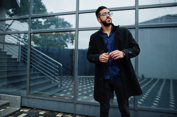 Middle eastern entrepreneur wear black coat and blue shirt, eyeglasses against office building smoking cigarette. - 写真・画像