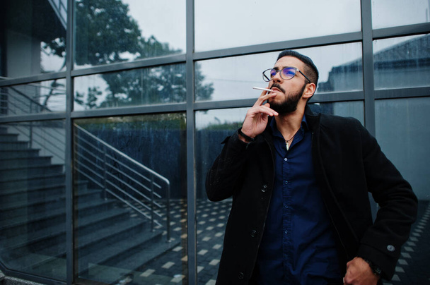 Middle eastern entrepreneur wear black coat and blue shirt, eyeglasses against office building smoking cigarette. - Zdjęcie, obraz