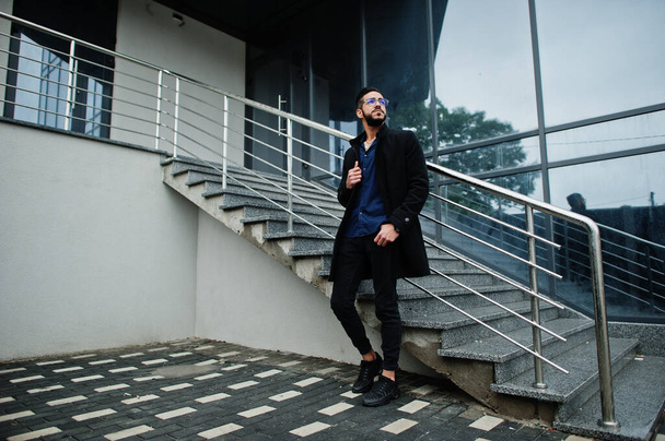 Imprenditore mediorientale indossare cappotto nero e camicia blu, occhiali contro edificio per uffici. - Foto, immagini