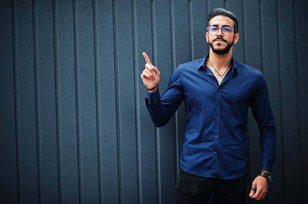 Middle eastern entrepreneur wear blue shirt, eyeglasses, against steel wall show finger up. - Φωτογραφία, εικόνα