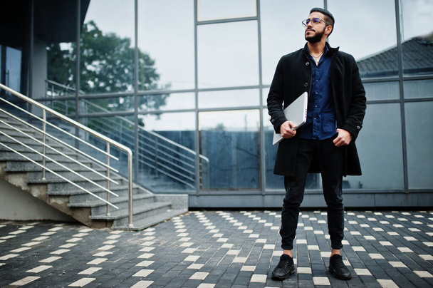 Middle eastern entrepreneur wear black coat and blue shirt, eyeglasses against office building with laptop. - Zdjęcie, obraz