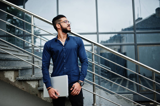 Middle eastern entrepreneur wear blue shirt, eyeglasses with laptop against office building. - Фото, зображення