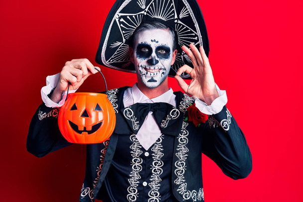 Joven hombre vistiendo el día mexicano del disfraz muerto sosteniendo calabaza haciendo signo de ok con los dedos, sonriendo gesto amistoso excelente símbolo  - Foto, imagen