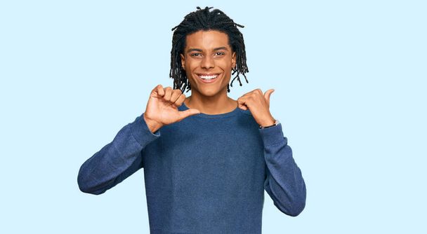 Молодой африканский американец в обычном зимнем свитере указывает на спину с поднятой рукой и большим пальцем, уверенно улыбаясь  - Фото, изображение