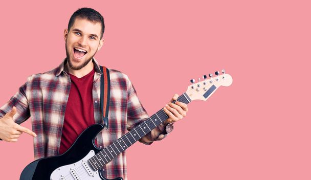 Fiatal jóképű férfi elektromos gitározik mosolyogva boldog mutogatás kézzel és ujjal  - Fotó, kép