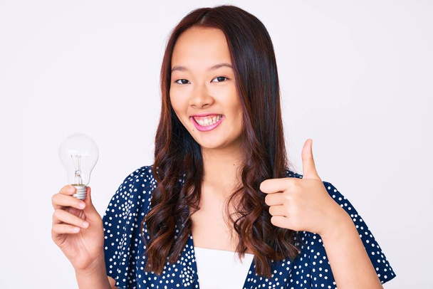 Jovem menina chinesa bonita segurando lâmpada para inspiração e ideia sorrindo feliz e positivo, polegar para cima fazendo excelente e sinal de aprovação  - Foto, Imagem