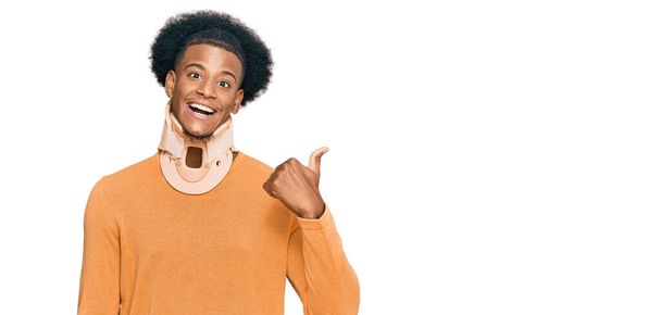 Africano americano com cabelo afro vestindo colar cervical pescoço sorrindo com o rosto feliz olhando e apontando para o lado com o polegar para cima.  - Foto, Imagem