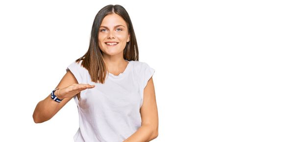 Junge kaukasische Frau in lässigem weißem T-Shirt gestikuliert mit Händen, die große und große Zeichen zeigen, Maßsymbol. Lächelnd in die Kamera blickend. Messkonzept.  - Foto, Bild