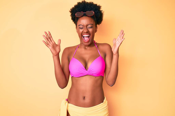 Mujer afroamericana joven vistiendo bikini celebrando loco y loco por el éxito con los brazos levantados y los ojos cerrados gritando emocionado. concepto ganador  - Foto, Imagen