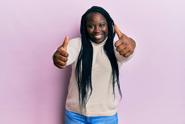 Fiatal fekete nő fonat visel alkalmi téli pulóver jóváhagyó csinál pozitív gesztus kézzel, hüvelykujj fel mosolyogva és boldog a siker. győztes gesztus.  - Fotó, kép