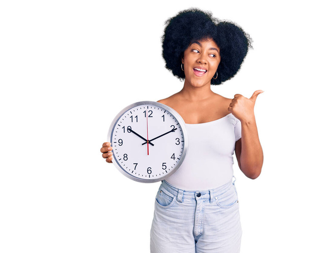 Büyük bir saat tutan Afrikalı Amerikalı genç bir kız başparmağıyla yan tarafa bakıyor ve ağzı açık gülümsüyor.  - Fotoğraf, Görsel