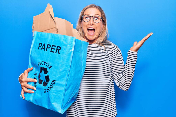 Mujer rubia caucásica de mediana edad sosteniendo papelera de reciclaje con papel y cartón celebrando la victoria con sonrisa feliz y expresión ganadora con las manos levantadas  - Foto, Imagen
