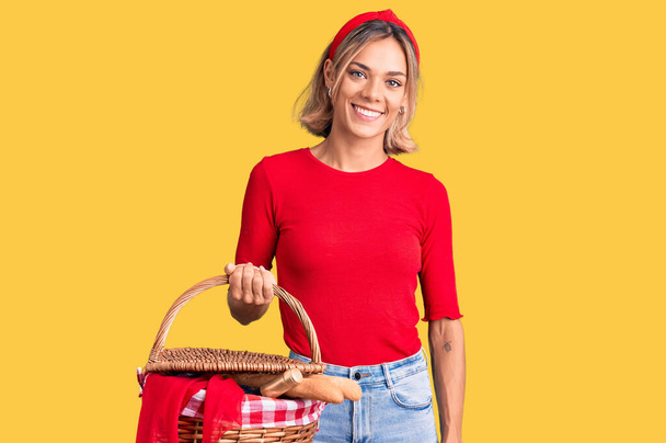 美しいcaucasian女性保持ピクニックウィッカーバスケットでパン見て正と幸せな立ちと笑顔で自信を持って笑顔を示す歯  - 写真・画像