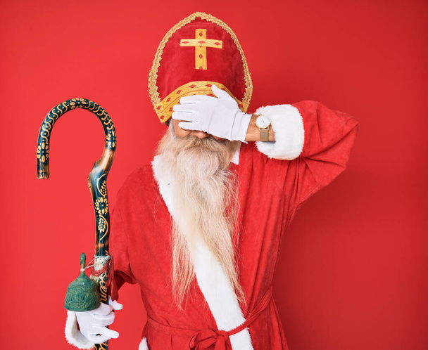 Gri saçlı ve uzun sakallı yaşlı bir adam geleneksel Aziz Nicholas kostümü giyiyor. Gözlerini eliyle kapatıyor, ciddi ve üzgün görünüyor. Görme, saklama ve reddetme konsepti  - Fotoğraf, Görsel