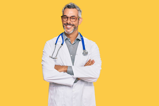Hombre de pelo gris de mediana edad vistiendo uniforme médico y estetoscopio cara feliz sonriendo con los brazos cruzados mirando a la cámara. persona positiva.  - Foto, Imagen