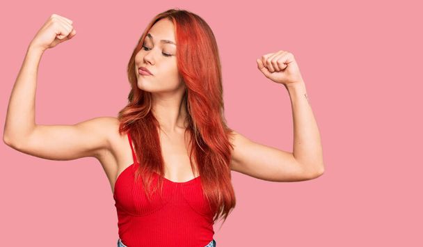 Genç kızıl saçlı kadın sıradan kıyafetler giyip gururla gülümseyen kol kaslarını gösteriyor. fitness konsepti.  - Fotoğraf, Görsel