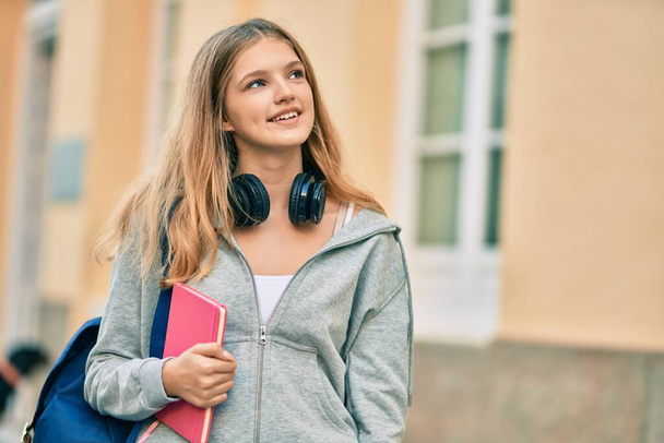 Bela adolescente estudante caucasiano usando fones de ouvido segurando livro na cidade. - Foto, Imagem