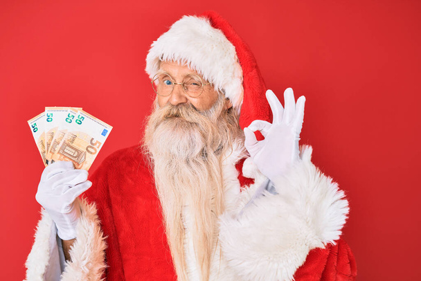 Staruszek w stroju Świętego Mikołaja trzyma euro robi znak ok palcami, uśmiechając przyjazny gest doskonały symbol  - Zdjęcie, obraz