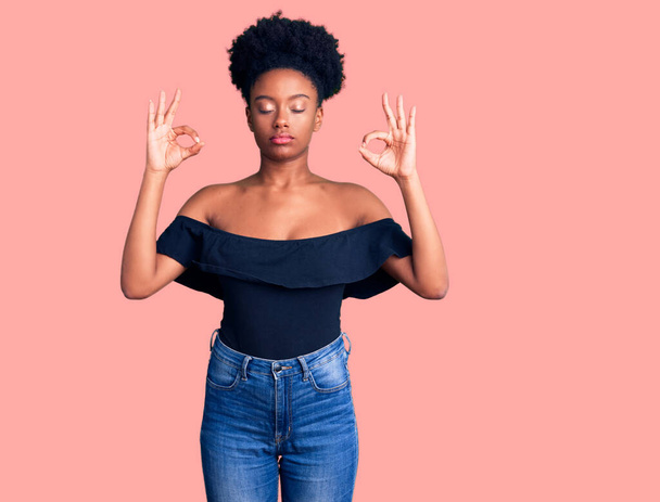 Mujer afroamericana joven con ropa casual relajarse y sonreír con los ojos cerrados haciendo gesto de meditación con los dedos. concepto de yoga.  - Foto, imagen