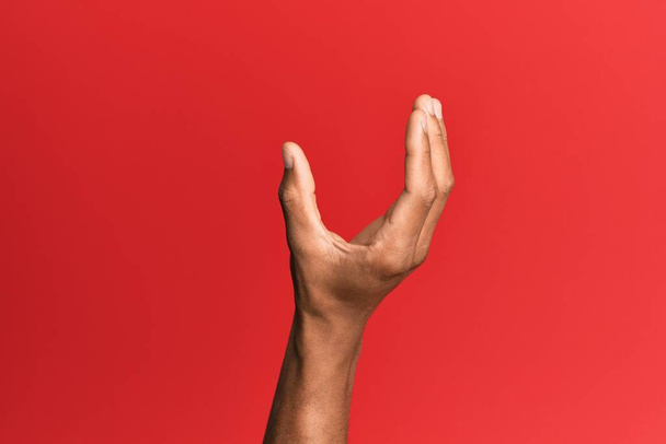 Χέρι του ισπανόφωνου ανθρώπου πάνω από το κόκκινο απομονωμένο φόντο μαζεύοντας και λαμβάνοντας αόρατο πράγμα, κρατώντας αντικείμενο με τα δάχτυλα που δείχνουν χώρο  - Φωτογραφία, εικόνα