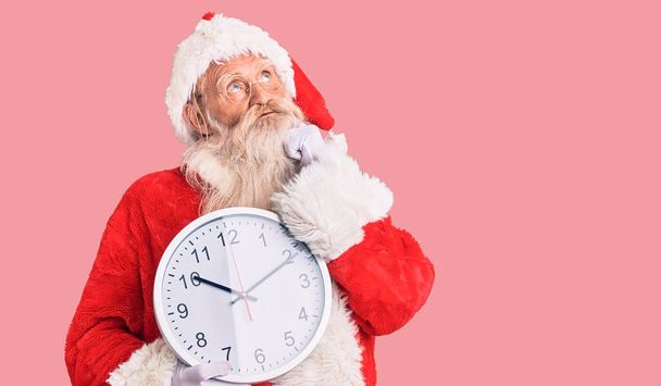 Old senior man with grey hair and long beard wearing santa claus costume holding clock serious face thinking about question with hand on chin, thoughtful about confusing idea  - Photo, Image