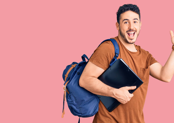 Sırt çantasında öğrenci çantası taşıyan İspanyol genç adam zaferi mutlu bir gülümseme ve ellerini kaldırarak kazanan ifadeyle kutluyor.  - Fotoğraf, Görsel
