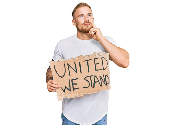 Young irish man holding united we stand banner serious face thinking about question with hand on chin, thoughtful about confusing idea  - Photo, Image