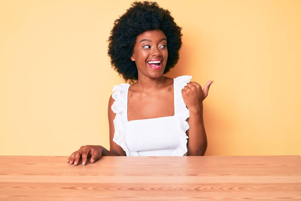 Joven mujer afroamericana vistiendo ropa casual sonriendo con la cara feliz mirando y señalando a un lado con el pulgar hacia arriba.  - Foto, imagen