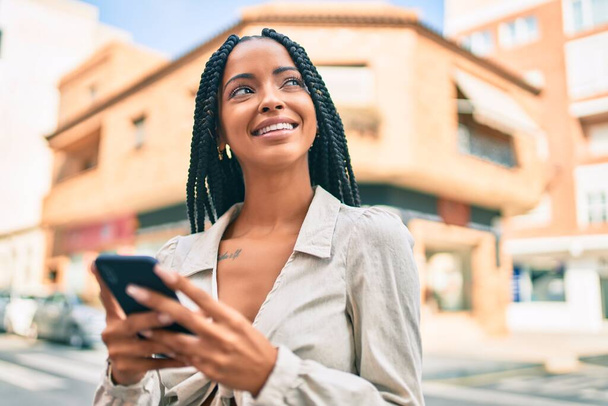 Молодая африканская американка улыбается счастливой, используя смартфон в городе. - Фото, изображение
