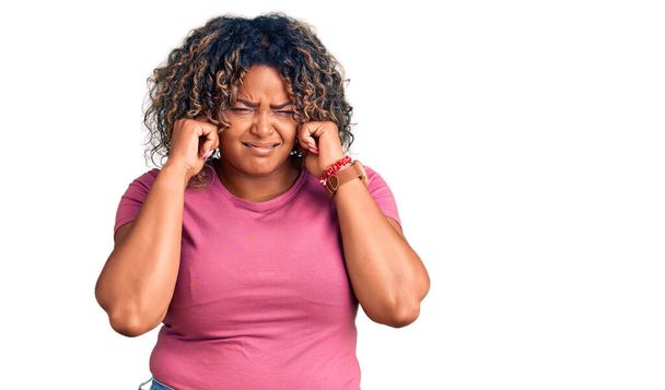 Jovem afro-americana plus size mulher vestindo roupas casuais cobrindo orelhas com dedos com expressão irritada para o ruído da música alta. conceito surdo.  - Foto, Imagem