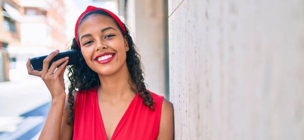 Chica afroamericana joven sonriendo feliz escuchando mensaje de audio utilizando el teléfono inteligente en la ciudad. - Foto, Imagen