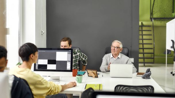 Idős férfi, vezető gyakornok hallgatja fiatal kollégáját, miközben laptopot használ, asztalnál ül, együtt dolgozik más alkalmazottakkal a modern irodában. - Fotó, kép