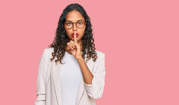 İş kıyafetleri giymiş genç Afro-Amerikalı kız dudaklarında parmaklarıyla sessiz olmak istiyor. sessizlik ve gizli konsept.  - Fotoğraf, Görsel