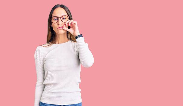 カジュアルな白いセーターとメガネの口と唇を身に着けている美しいブルネットの若い女性は指でzipとしてシャットダウンします。秘密裏に黙ってタブーを語る  - 写真・画像