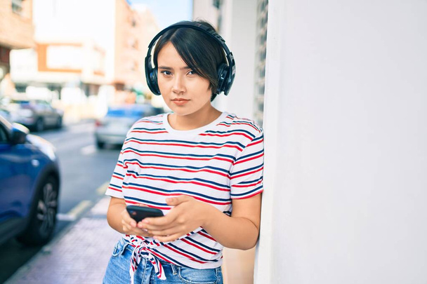Giovane ragazza latina con espressione grave utilizzando smartphone e cuffie in città - Foto, immagini
