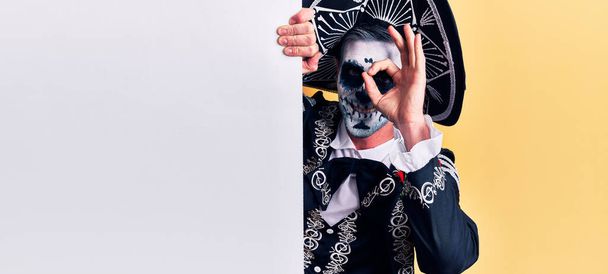 Молодой человек, одетый в мексиканский день мертвого костюма, держит пустой баннер, улыбаясь счастливо, делая хорошо знак с рукой на глазу, глядя сквозь пальцы  - Фото, изображение