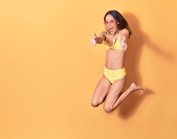 Young beautiful girl wearing bikini and sunglasses smiling happy. Jumping with smile on face doing ok sign with thumbs up over isolated yellow background - Foto, Imagem