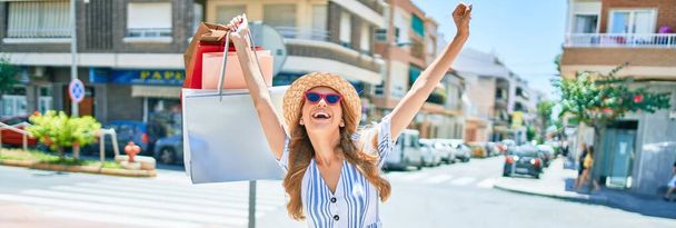 Νεαρή όμορφη πωλήτρια γυναίκα χαμογελά ευτυχισμένη πηγαίνοντας στα καταστήματα πωλήσεις κρατώντας τσάντες ψώνια ourtdoors, χαμογελώντας ευτυχισμένη με ανοιχτές αγκάλες απολαμβάνοντας τον ήλιο - Φωτογραφία, εικόνα