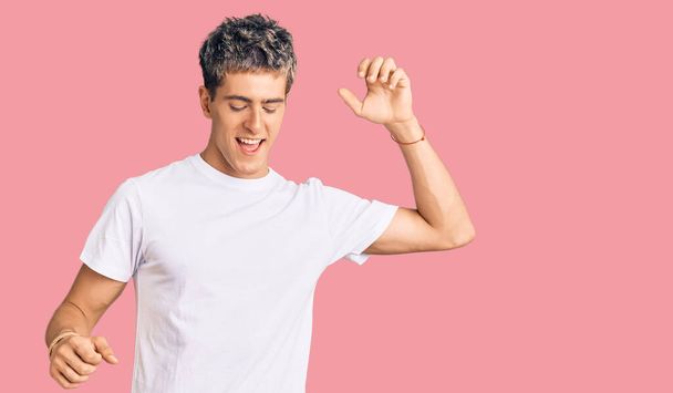 Junger gutaussehender Mann in lässigem weißem T-Shirt tanzt fröhlich und fröhlich, lächelnd bewegt er sich lässig und selbstbewusst und hört Musik  - Foto, Bild