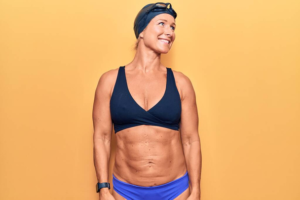 Middle age caucasian blonde woman wearing bikini,  swimming cap and diving googles looking away to side with smile on face, natural expression. laughing confident.  - Photo, Image