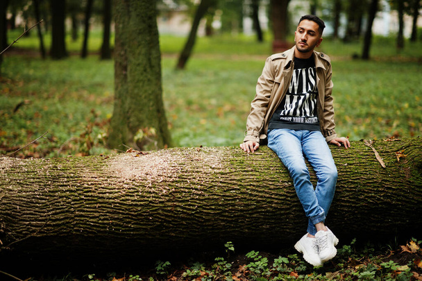 Stylish kuwaiti man at trench coat sit on tree at wood. - Fotoğraf, Görsel