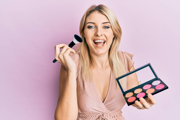 Young caucasian woman holding makeup brush and blush palette smiling and laughing hard out loud because funny crazy joke.  - Photo, Image